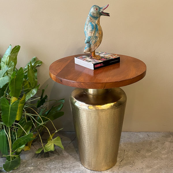 A round wooden top side table with a hammered metallic gold cylindrical pedestal base, styled with a decorative bird figurine and a book, set against a textured beige wall with a green plant on the left.