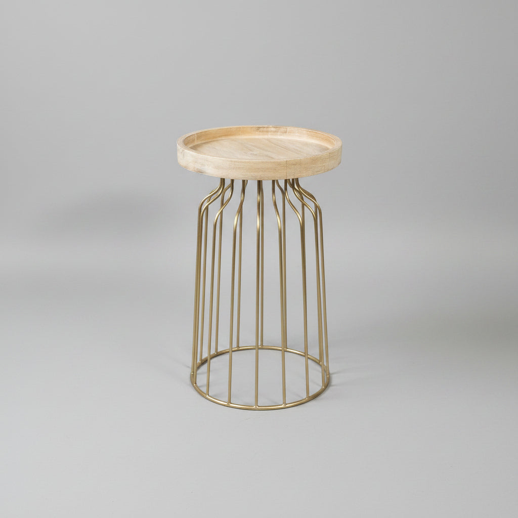 A round wooden tray top side table with a gold metal wire cage base, set against a textured beige wall and gray stone floor with a green plant on the left.